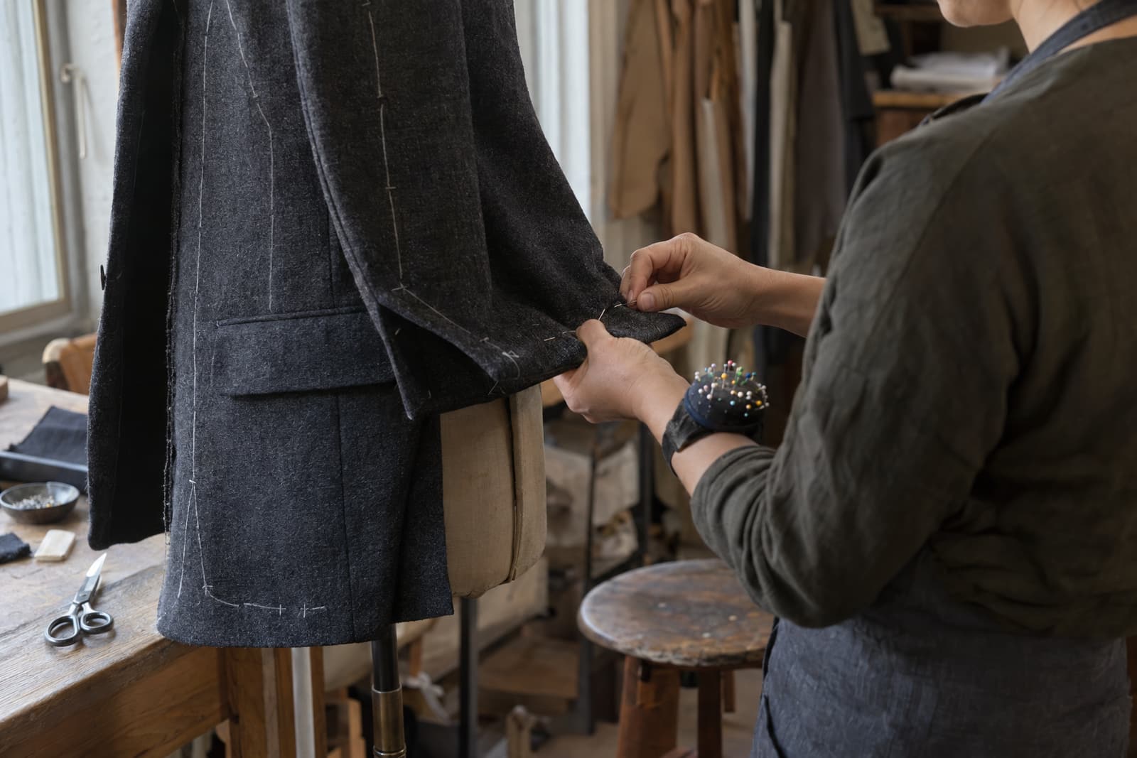 Two hands pin the sleeve of a charcoal wool jacket on a dress form, next to a wooden workbench with scissors and a dish of pins, while the person wears a pin cushion on the wrist.