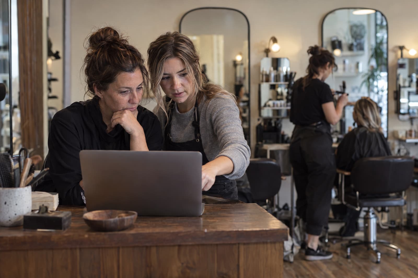 Two women sit close together at a wooden desk looking at a laptop whose screen faces away, one pointing at it, while in the background a hairdresser works on a seated person in front of tall arched mirrors.