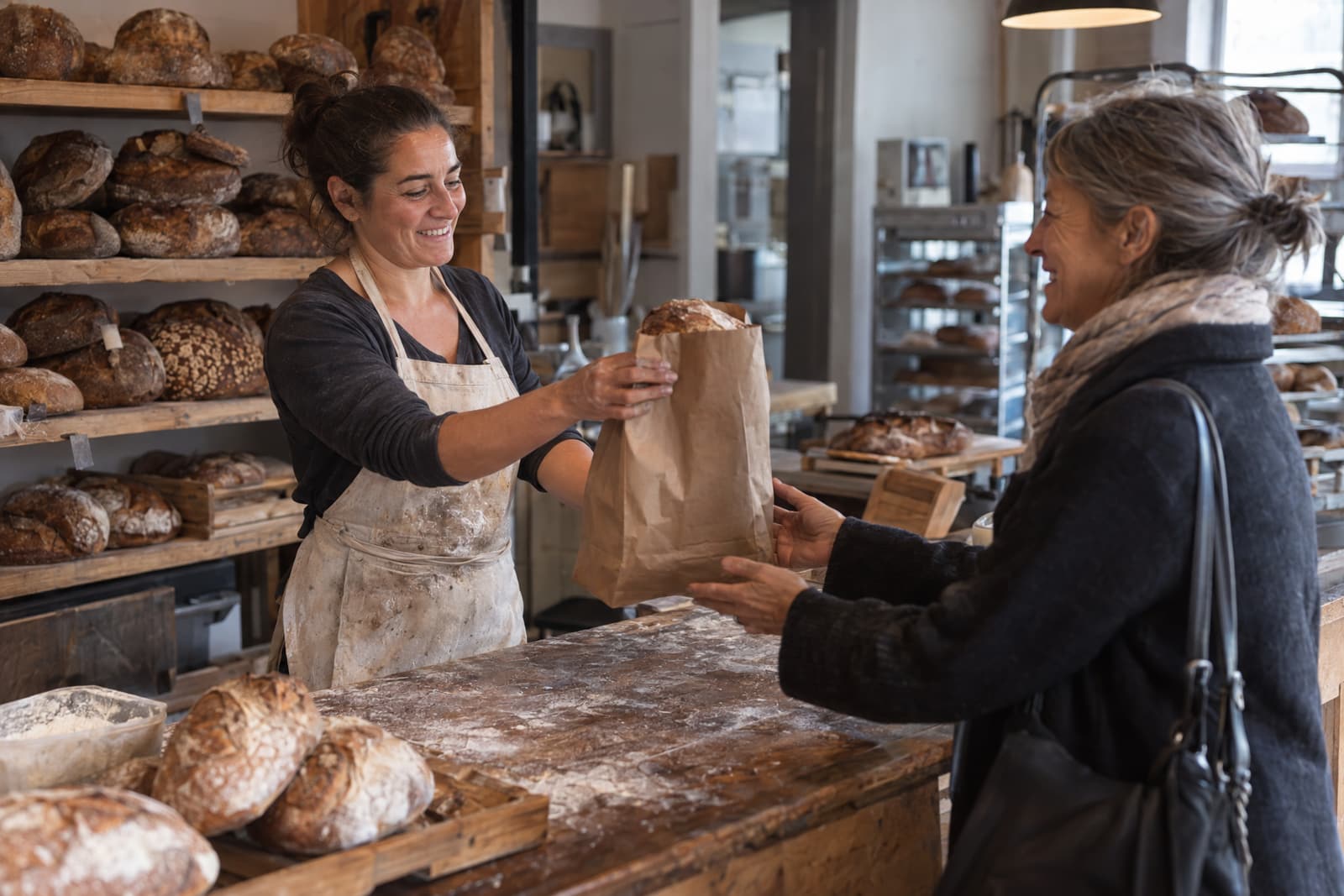 A smiling woman in a flour-dusted apron hands a paper bag with a loaf of bread across a floured wooden counter to a smiling grey-haired woman in a dark coat and scarf, with shelves of round loaves behind them.