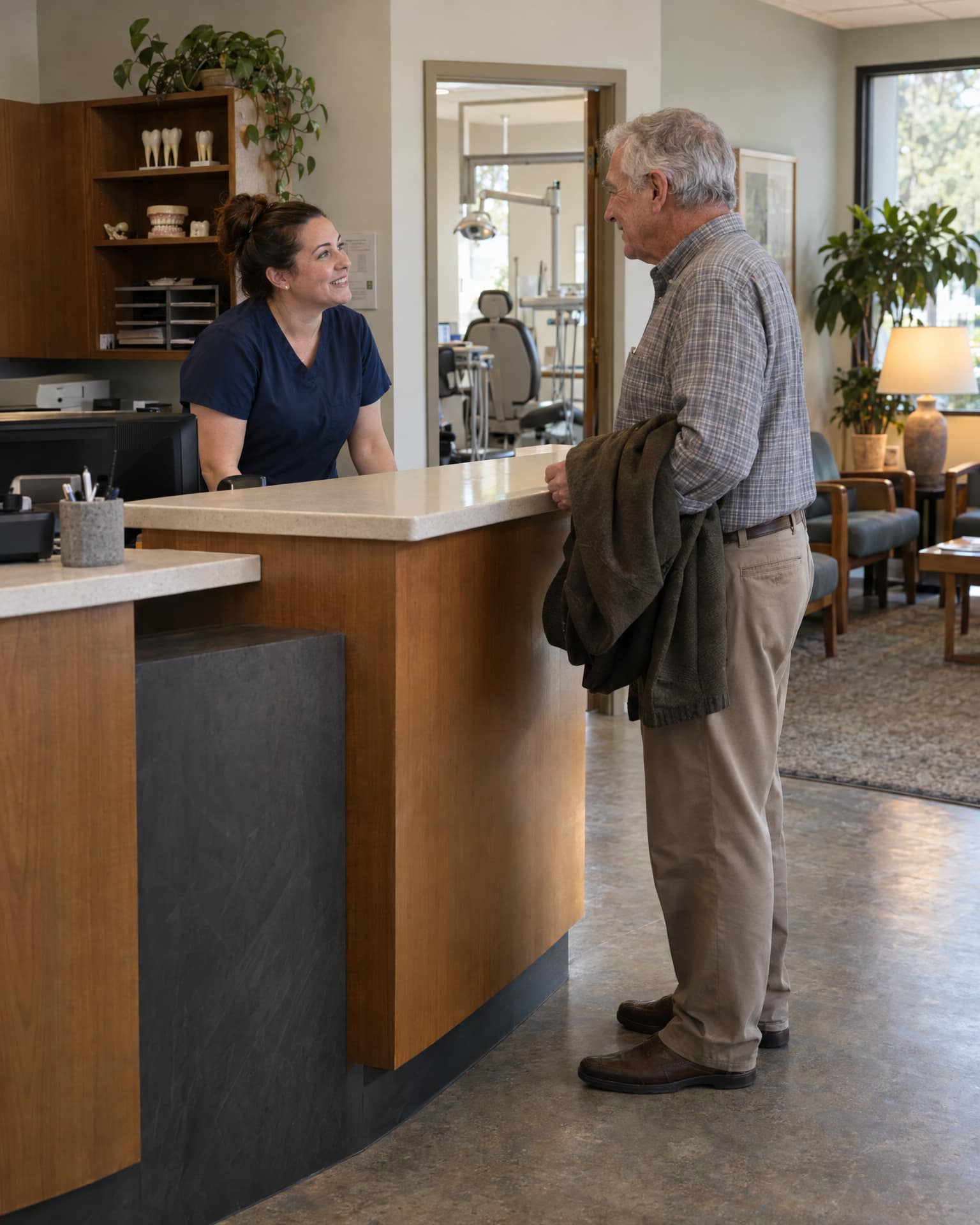A smiling woman in navy scrubs stands behind a wooden reception counter talking with an older man in a plaid shirt and khaki trousers who holds a dark jacket over his arm, with tooth models on a shelf, a dental chair visible through a doorway, and a waiting area with plants and a lamp to the right.