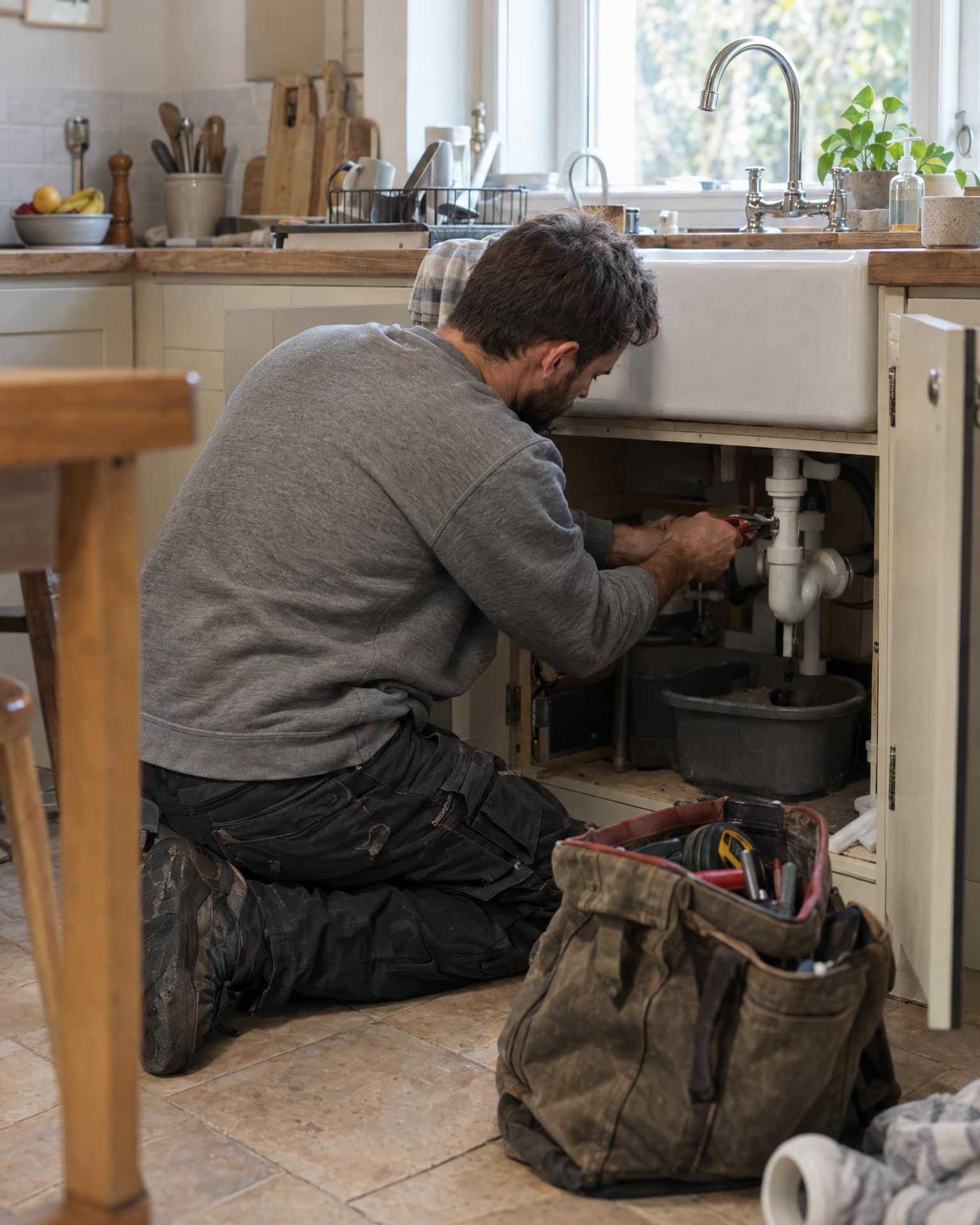 A bearded man in a grey sweatshirt and dark work trousers kneels on a tiled kitchen floor, working with pliers on the white pipes under a ceramic sink, beside an open canvas tool bag.