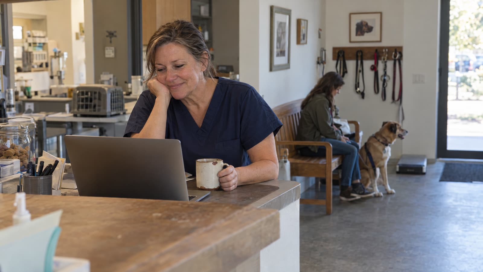 A woman in navy scrubs leans on a wooden reception counter, chin resting on one hand, smiling at a laptop while holding a ceramic mug, while behind her a woman sits on a wooden bench beside a seated dog under a row of leashes hanging on wall hooks.