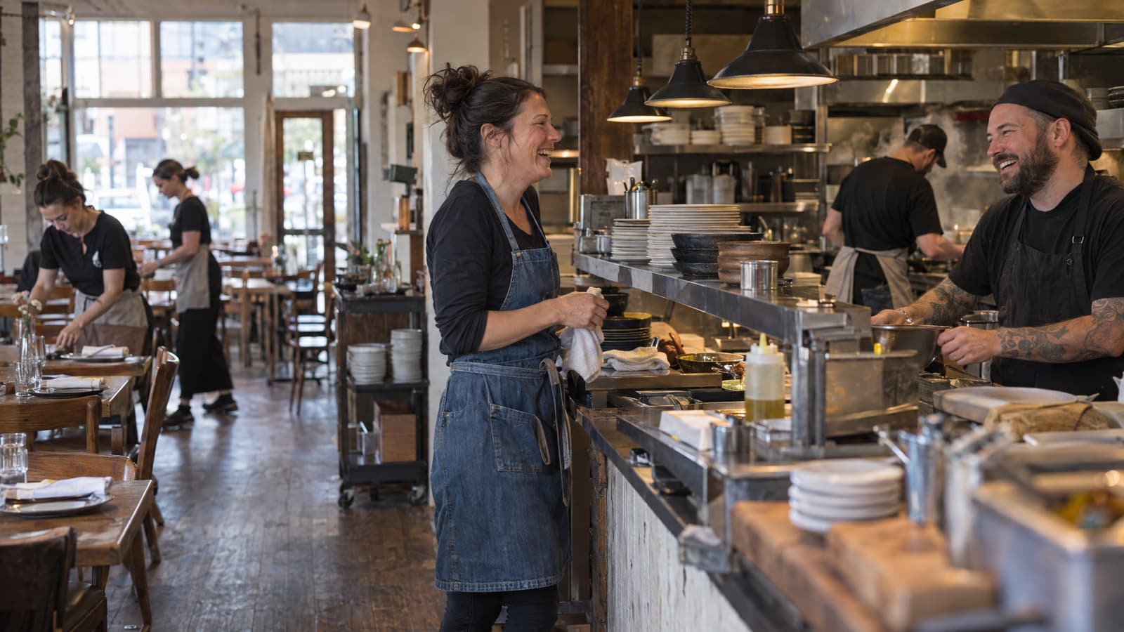 A woman in a denim apron holding a white towel laughs with a bearded, tattooed cook in a black cap across a steel counter in a restaurant, while two women in aprons set wooden tables by large front windows and another cook works at the stove behind him.