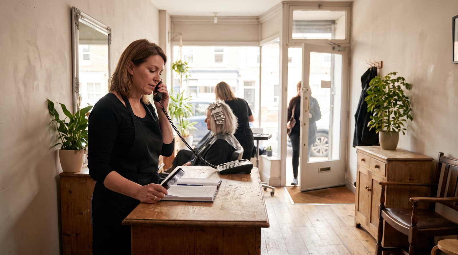 A salon receptionist on a corded phone writes in an appointment book while a woman in hair foils waits behind her.