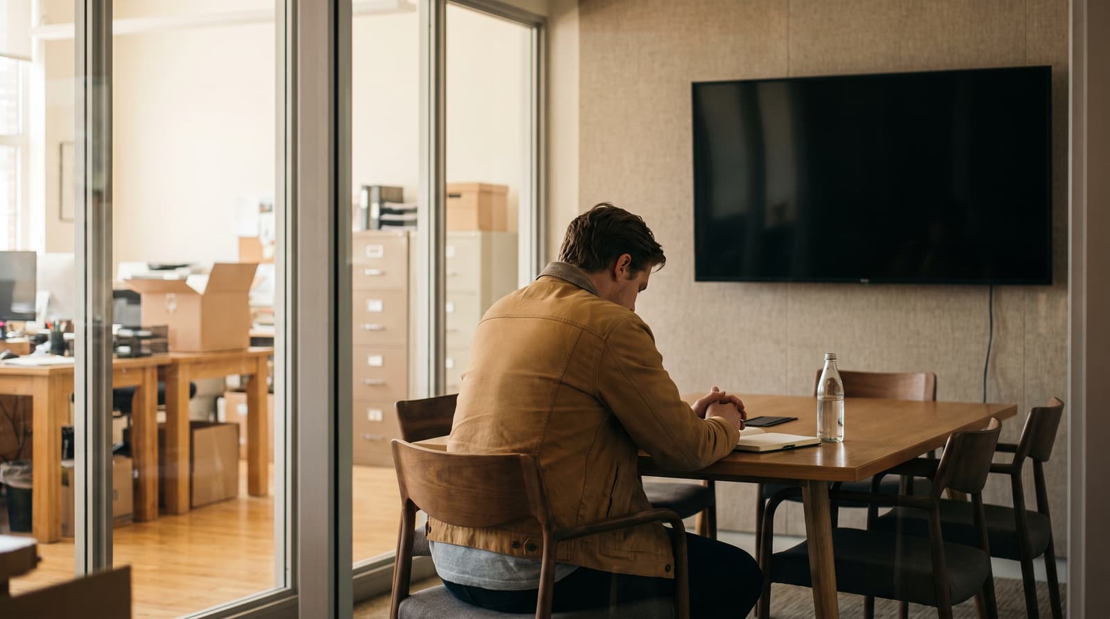 A man in a brown jacket sits alone at a wooden meeting table with clasped hands, a notepad and a water bottle beneath a blank wall-mounted screen.