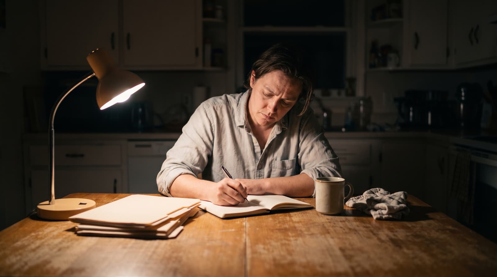 A woman writes in an open notebook at a wooden kitchen table, with a lit desk lamp, stacked paper folders and a mug beside her and dark kitchen cabinets behind.