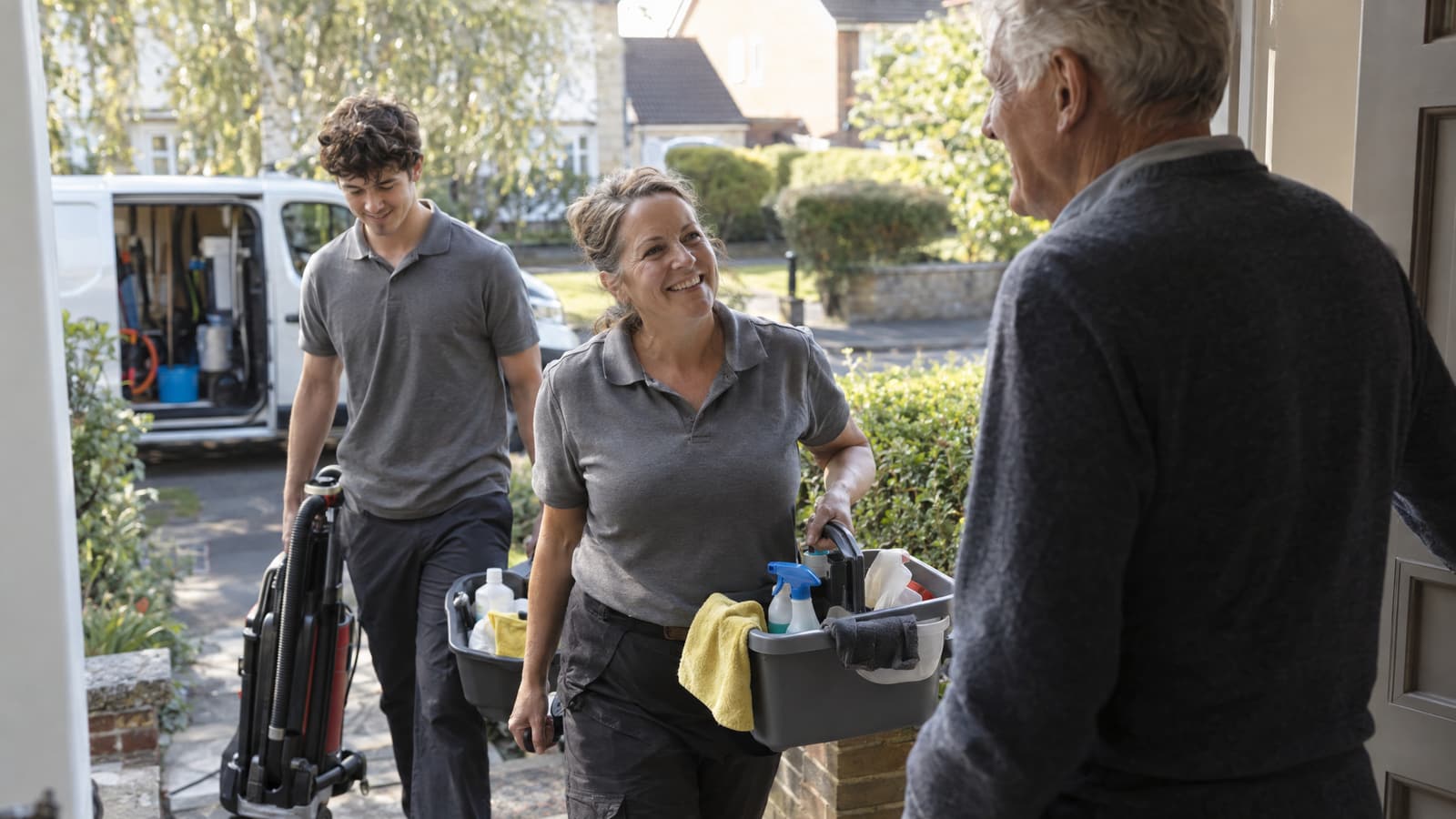 A smiling woman carrying grey caddies of cleaning bottles and cloths and a young man wheeling an upright vacuum walk up a garden path toward an older man standing at an open front door, with a white van parked on the street behind them.