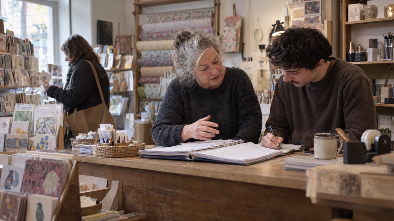 An older woman with grey hair in a bun gestures with one hand over an open ring binder on a wooden shop counter while a young man beside her writes in a notebook, with shelves of cards and rolled paper behind them and a woman browsing cards at the left.