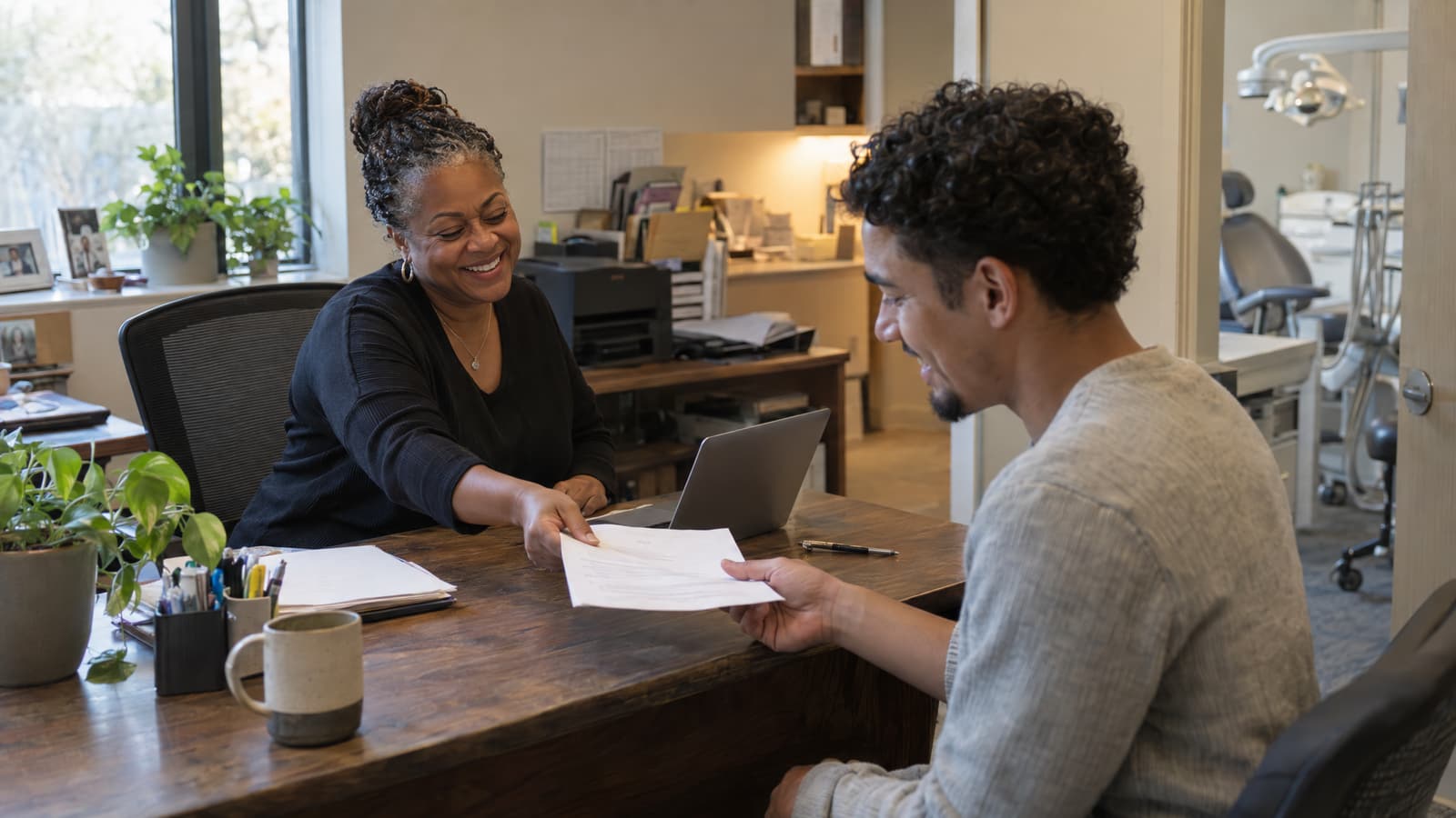 A smiling woman in a black sweater hands a sheet of paper across a wooden desk to a smiling young man in a grey sweater, in an office with a window, plants, a mug and a laptop, with a dental chair and lamp visible through a doorway on the right.