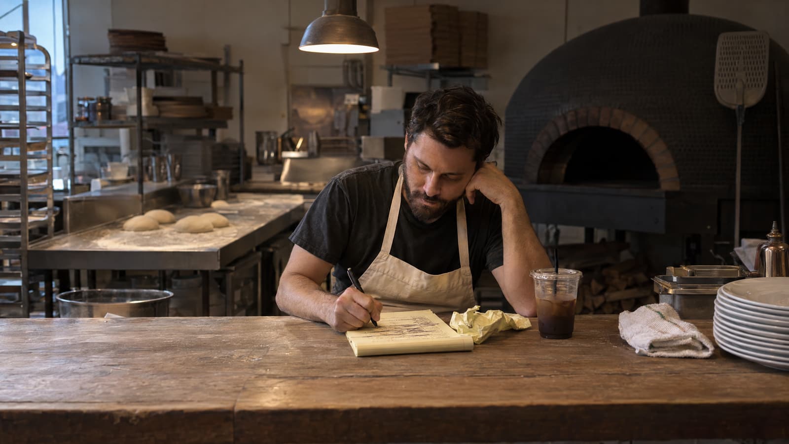 A bearded man in a dark T-shirt and canvas apron sits at a worn wooden counter writing on a yellow legal pad, resting his head on one hand, with a crumpled sheet, an iced coffee and a stack of plates beside him and a dough bench and domed brick oven behind him.