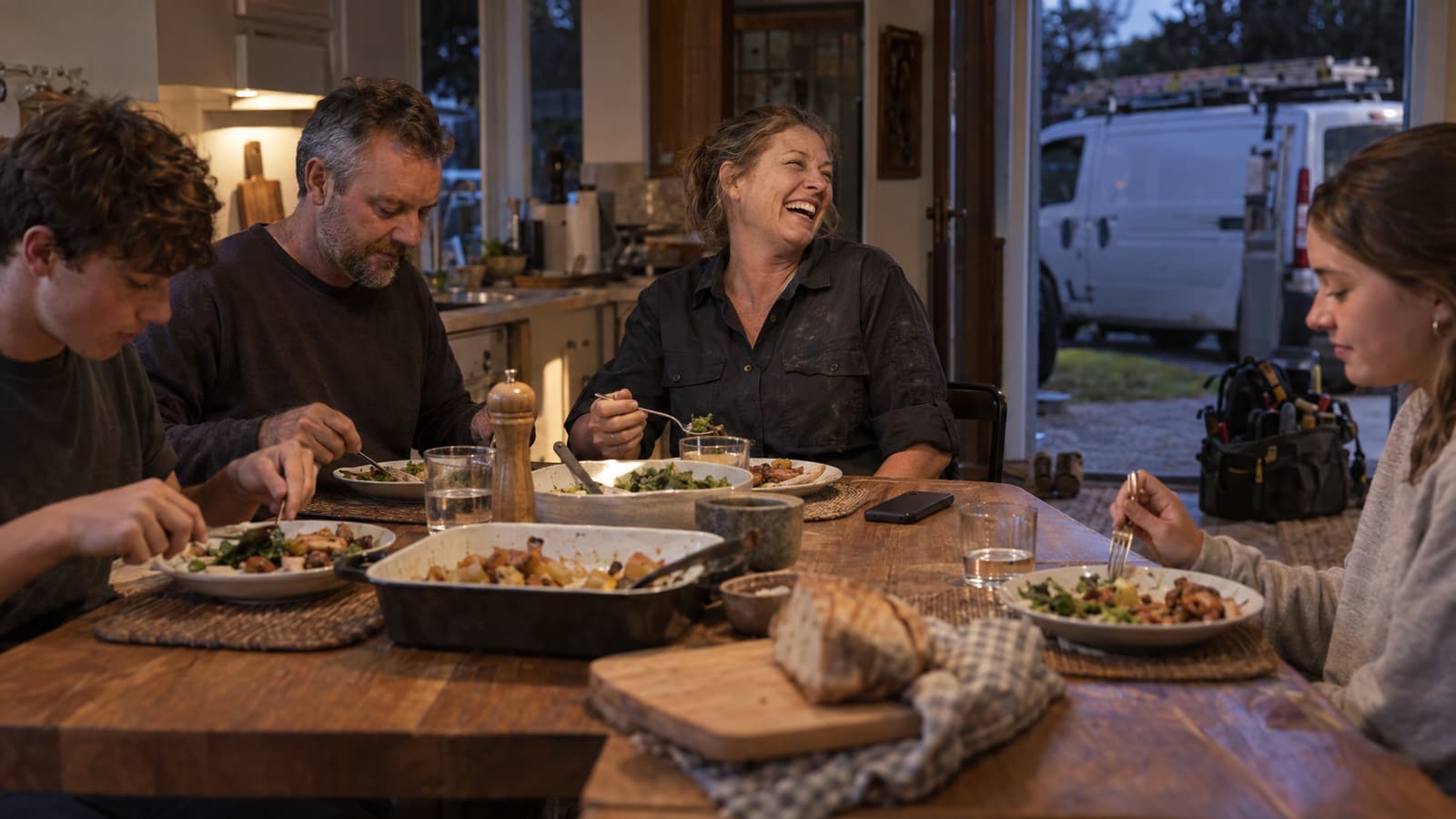 A woman in a dusty dark work shirt laughs at a wooden kitchen table while a man and two teenagers eat dinner around her, with a phone lying on the table and a white van with a roof ladder rack parked outside the open door at dusk.