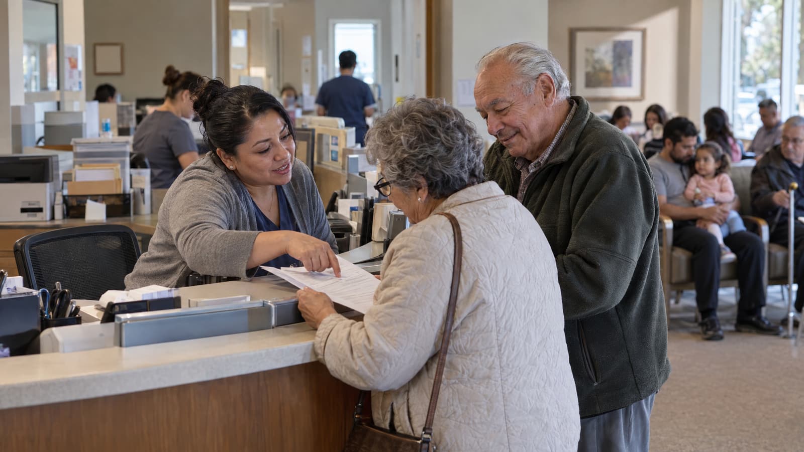 A woman behind a clinic front desk leans forward and points to a sheet of paper held by an older woman in a quilted jacket, while an older man in a dark green fleece looks on smiling and people sit in a waiting area behind them.