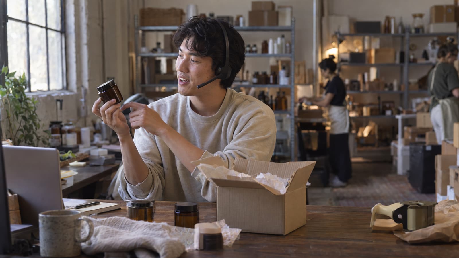 A young man wearing a headset with a microphone sits at a wooden table, holding up a small amber glass jar and pointing at it. In front of him are an open cardboard box with tissue paper, more amber jars and a mug, and behind him people work among shelves of bottles and boxes beside a large window.