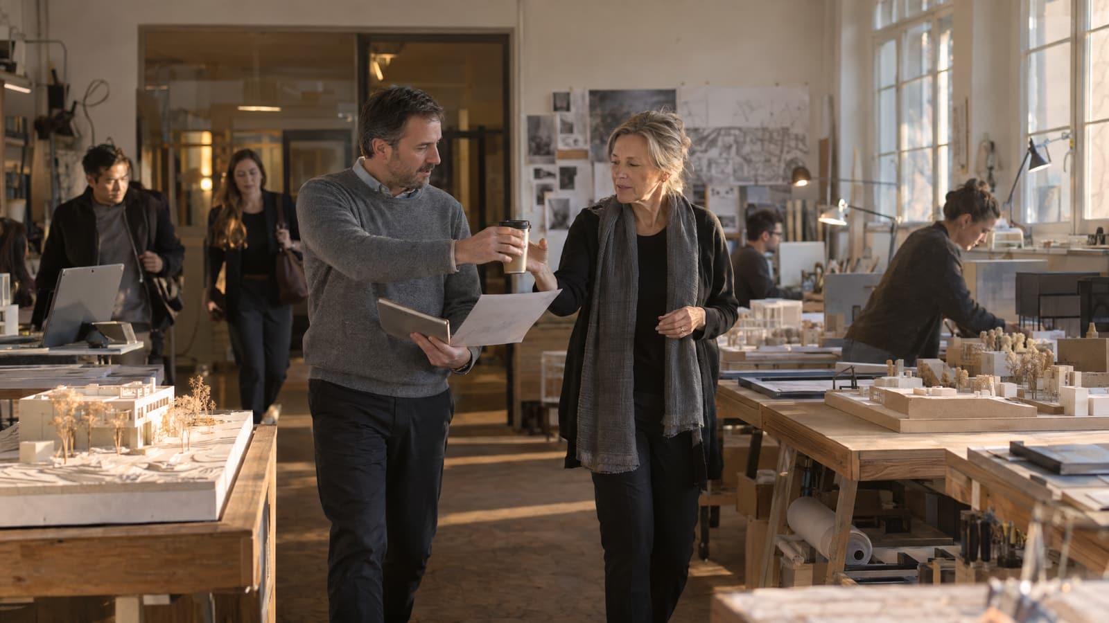 A man in a grey sweater holding a tablet and a sheet of paper hands a paper coffee cup to a woman in a black cardigan and grey scarf as they walk between wooden tables of white architectural models in a sunlit studio, with other people working and arriving behind them.
