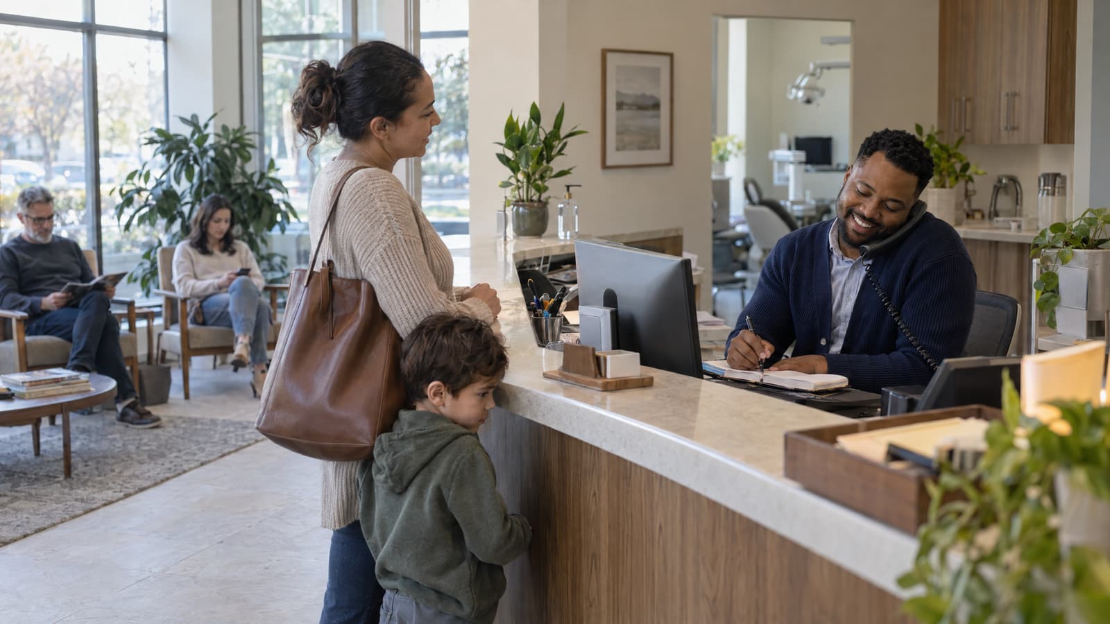 A smiling man in a navy cardigan holds a desk phone against his shoulder and writes in a paper book at a clinic front desk, while a woman with a leather bag and a small boy stand at the counter and two people sit in a bright waiting room behind them.