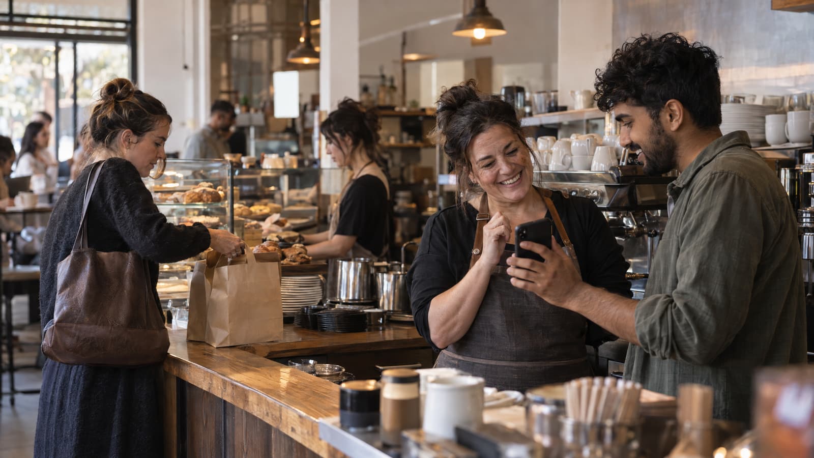In a busy café, a smiling woman in a dark apron behind a wooden counter looks at a phone held up by a bearded young man in a green shirt, while another woman at the left takes something from a brown paper bag in front of a pastry case.