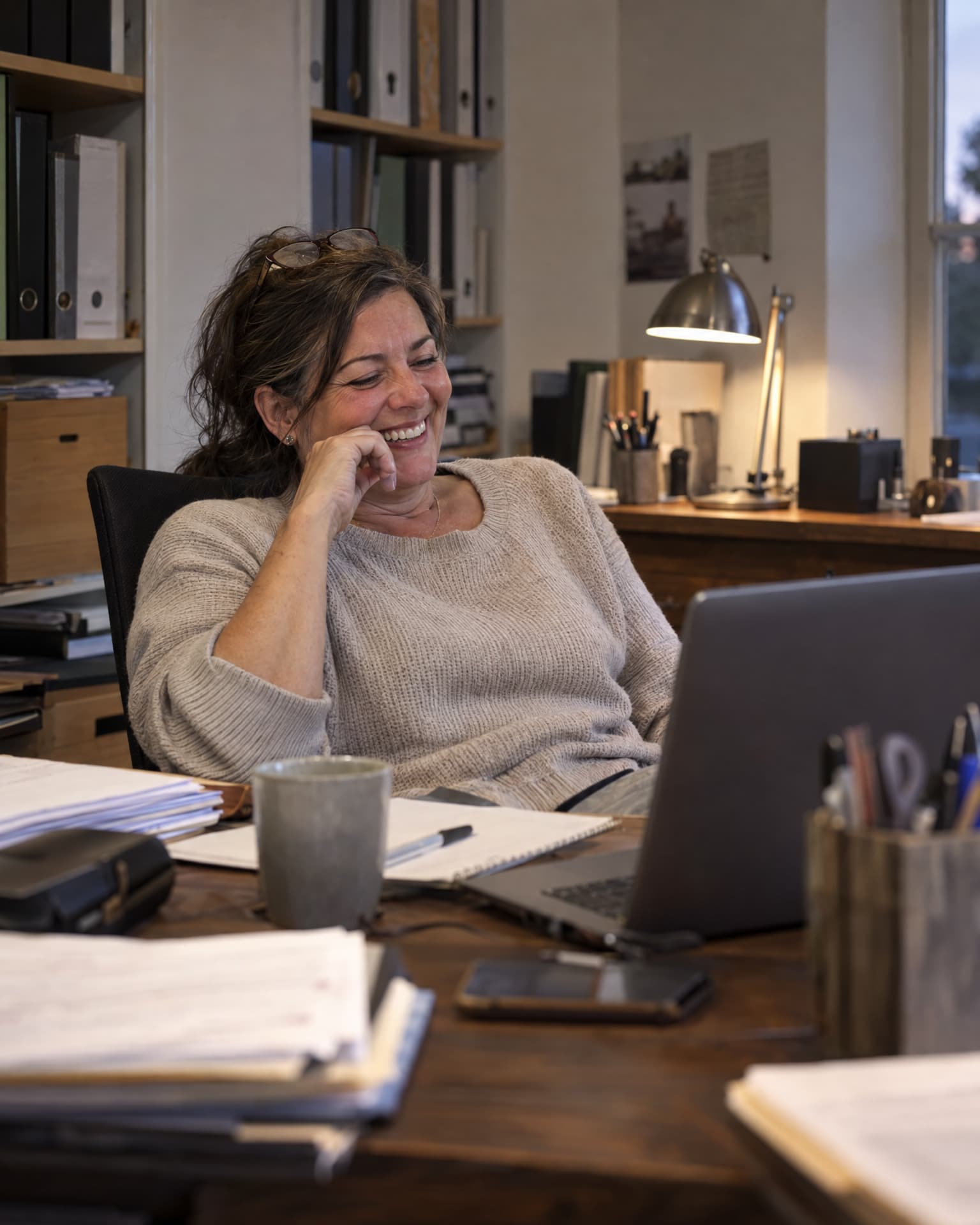 A woman with glasses pushed up into her dark hair leans back in an office chair, smiling with her hand at her chin as she looks at a laptop on a wooden desk covered with papers, a mug, a phone and a pen pot, with binders on shelves and a lit desk lamp behind her.