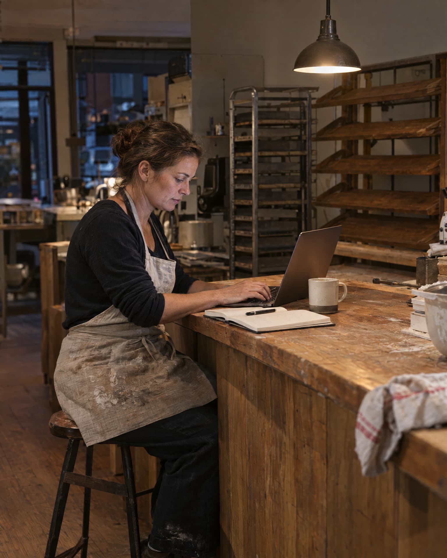 A woman in a dusty linen apron sits on a wooden stool at a long worn wooden counter typing on a laptop, with a notebook, pen and mug beside her, empty wooden bread racks and a metal tray rack behind her under a single lit pendant lamp.